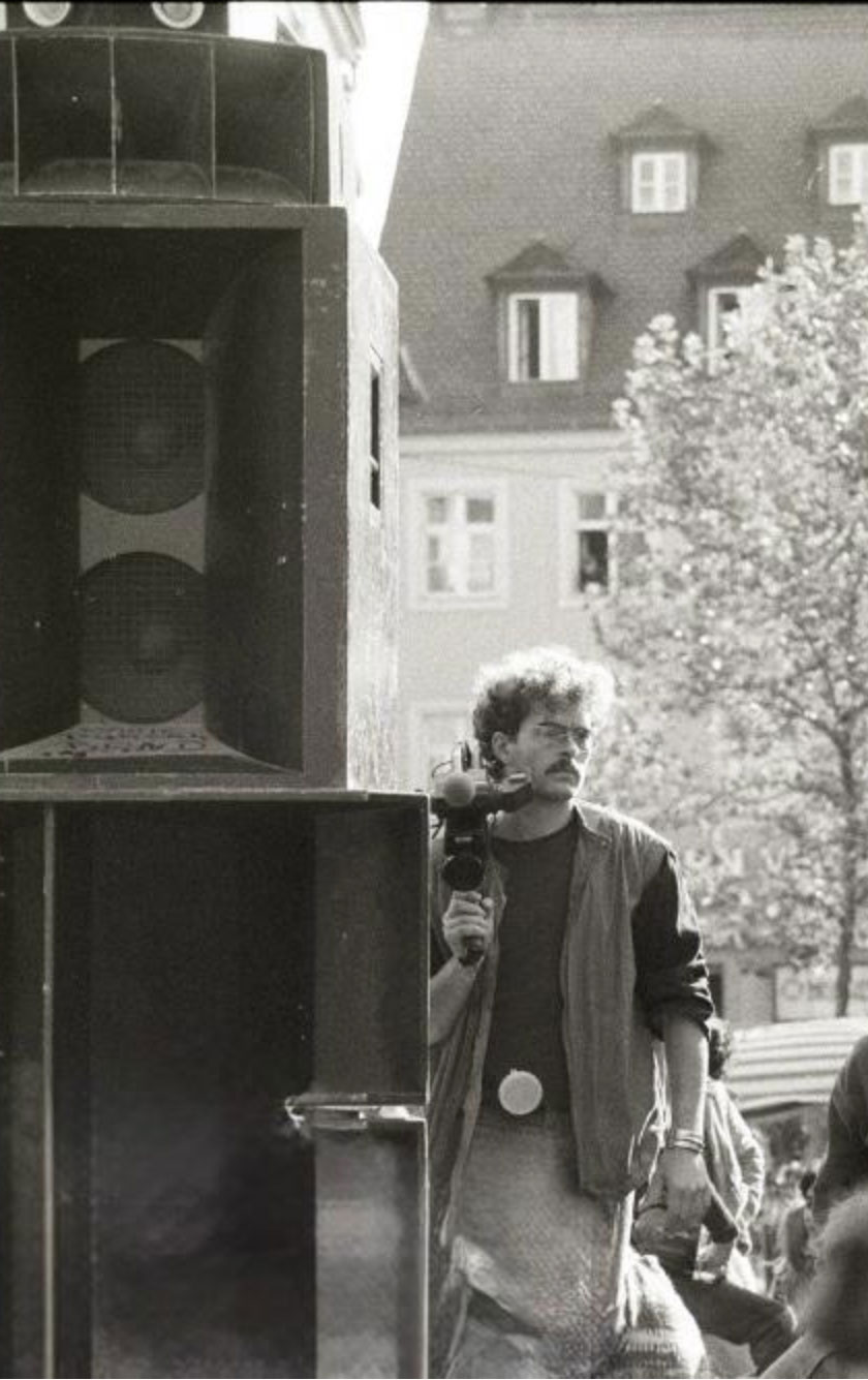 Ralph Kloos in jungen Jahren mit Videokamera auf der Schulter, neben ihm sehr großen Lautsprecher und schemenhaft ist Publikum zu sehen.